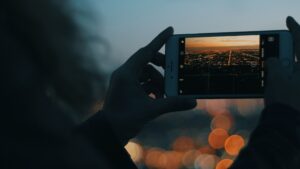 Ein smartphone wird quer von einer Hand gehalten. Es wird eine städtische Landschaft fotografiert. Man sieht verschwommen warm-goldene Lichter vom Sonnenuntergang und der Stadt in der Ferne. Außerdem sieht man einen Teil des Himmels, der in einem schwachen verwaschenem Blau einen Kontrast zu den Lichtern bildet.