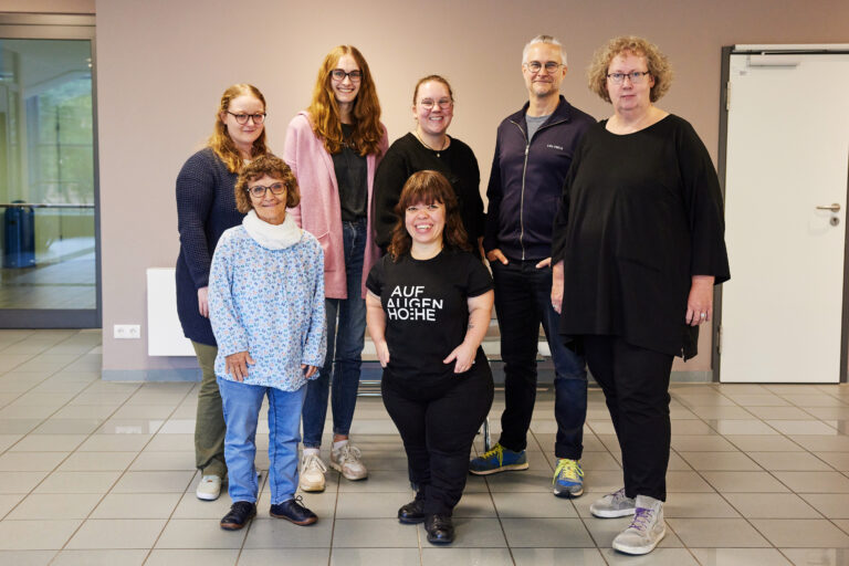 Gruppenbild der stehenden Workshop-Teilnehmenden mit der kleinwüchsigen Fotografin Anna Spindelndreier im Vordergrund.