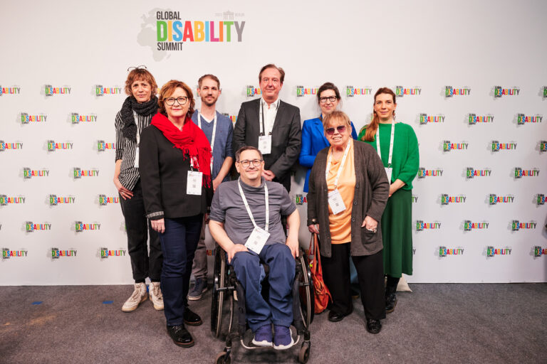 Vor einer weißen „Pressewand“, die mehrfach mit dem bunten Logo des „Global Disability Summits“ bedruckt ist, haben sich acht Menschen mittleren Alters zum Gruppenfoto aufgestellt, die in schicker Kleidung freundlich in die Kamera lächeln. Hinter einem Rollstuhlfahrer, der jeweils links und rechts von einer Frau flankiert wird, stehen in der hinteren Reihe eine weitere Frau, zwei Männern und noch mal zwei Frauen. Bei der zweiten von rechts handelt es sich um LAG-Geschäftsführerin Melanie Ahlke, die einen samtblauen Blazer, eine schwarze Hornbrille und die langen, dunklen Haare gescheitelt zu einem Zopf trägt.
