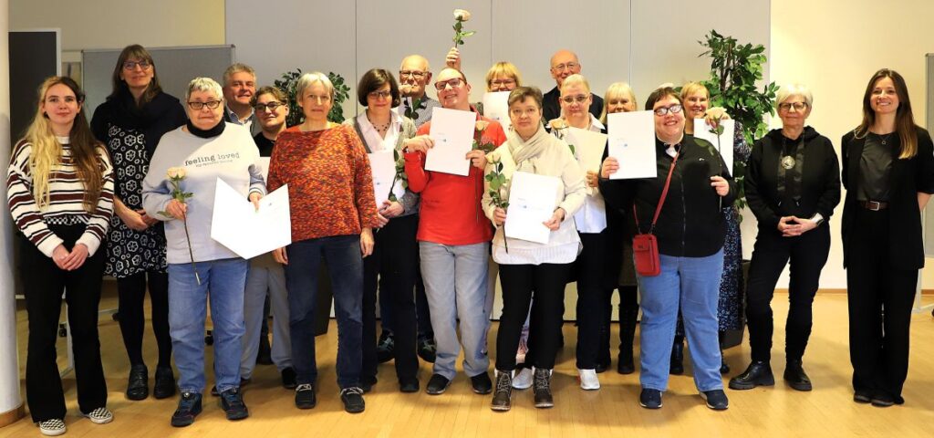 Rund 20 Personen haben sich vor einer grauen Wand zum Gruppenbild aufgestellt. Die Teilnehmenden des HospInk-Kurses halten stolz ihr Abschlusszeugnisse und eine beige Rose in den Händen. Unter ihnen sind einige Männer, vor allem aber Frauen unterschiedlicher Altersgruppen und in farbenfroher Kleidung, sodass ein bunter, fröhlicher. vielfältiger Eindruck entsteht.