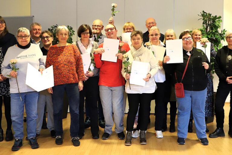 Rund 20 Personen haben sich vor einer grauen Wand zum Gruppenbild aufgestellt. Die Teilnehmenden des HospInk-Kurses halten stolz ihr Abschlusszeugnisse und eine beige Rose in den Händen. Unter ihnen sind einige Männer, vor allem aber Frauen unterschiedlicher Altersgruppen und in farbenfroher Kleidung, sodass ein bunter, fröhlicher. vielfältiger Eindruck entsteht.