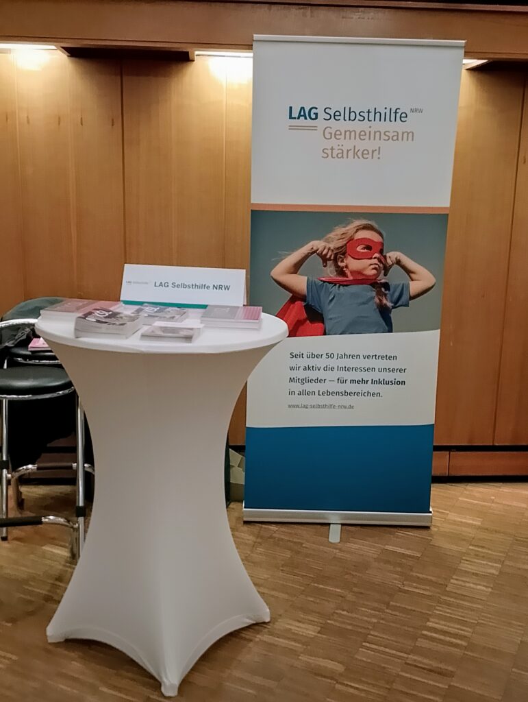 Auf einem Stehtisch liegen verschiedene Broschüren und das Namensschild der LAG Selbsthilfe NRW liegen. Dahinter ist unser Roll-Up zu sehen, das passend zum Motto „Gemeinsam stärker“ ein Mädchen im Superheld*innen-Umhang und folgenden Text zeigt: Seit über 50 Jahren vertreten wir aktiv die Interessen unserer Mitglieder – für mehr Inklusion in allen Lebensbereichen.