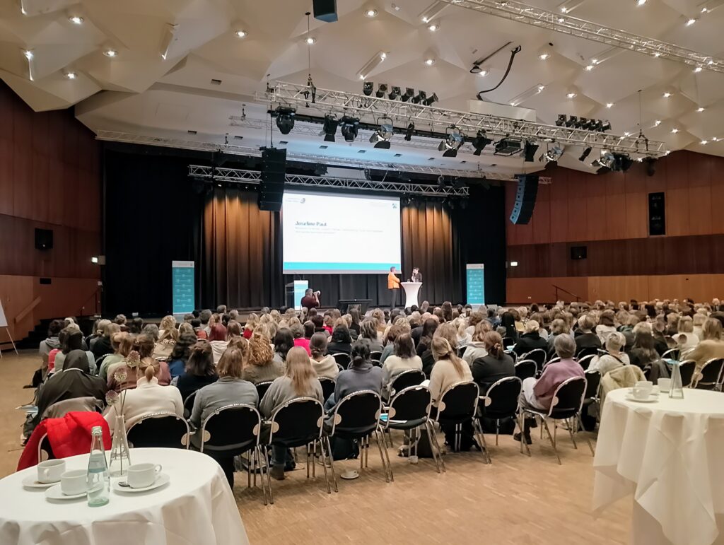 Blick von hinten über das zahlreich versammelte Publikum im Veranstaltungssaal, das von vollen Stuhlreihen zum leicht erhöhten Podium hochschaut. Dort unterhalten sich gerade Josefine Paul und die Moderatorin an einem Stehtisch. Hinter ihnen ist die Präsentation mit dem Namen der Ministerin auf eine Leinwand geworfen.