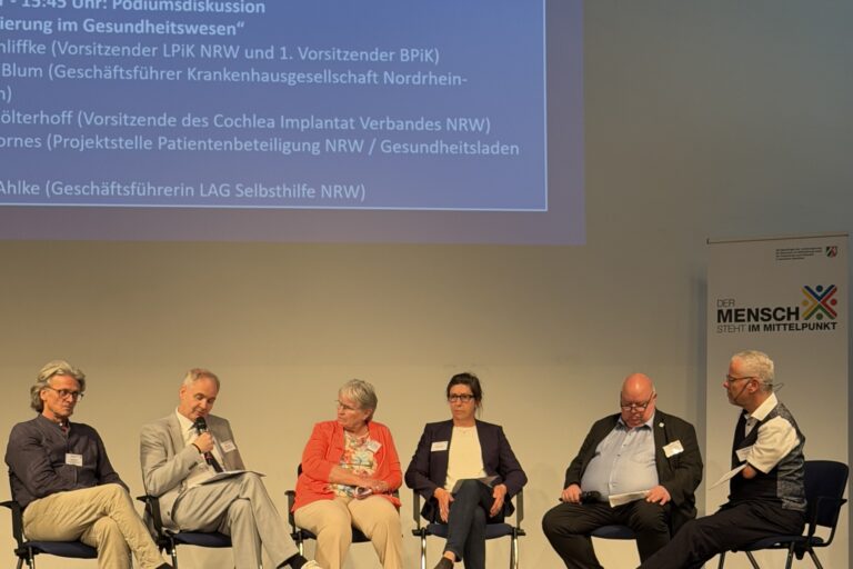 Auf einem Podium sitzen 6 Menschen auf Stühlen in einem Halbkreis - v. l. n. r.: Ein Mann mit mittellangen, grau gewellten Haaren und randloser Brille in dunkelblauem Hemd, beiger Hose und weinroten Sneakern. Ein älterer Herr mit kurzem Haar in grauem Anzug, der in das Mikro in seiner Hand spricht und dabei auf die Zettel auf seinem Knie schaut. Eine ältere Frau grauen, mittellangen Haaren in einer orangefarbenen Strickjacke über einem bunt gemustertem Shirt, die zum Sprecher neben ihr schaut. LAG-Geschäftsführerin Melanie Ahlke mit langen, dunklen, hochgesteckten Haaren und Hornbrille, in dunkler Hose und Blazer über einem hellem Shirt. Ein korpulenterer Mann mit Halbglatze, Brille und hellblauem Hemd unter einem dunklem Anzug. Und Moderator Rainer Schmidt im seitlichen Profil mit kurzen, grauen Haaren, Headset, Brille, dunkler Hose, Weste und weißem, kurzärmeligem Hemd, unter dem der linke Armstumpf hervorguckt. Im Hintergrundsind außerdem ein Roll-Up mit der Aufschrift „Der Mensch steht im Mittelpunkt“ sowie die Projektion einer Präsentation mit Ausschnitten der Agenda zur Podiumsdiskussion zu erahnen.
