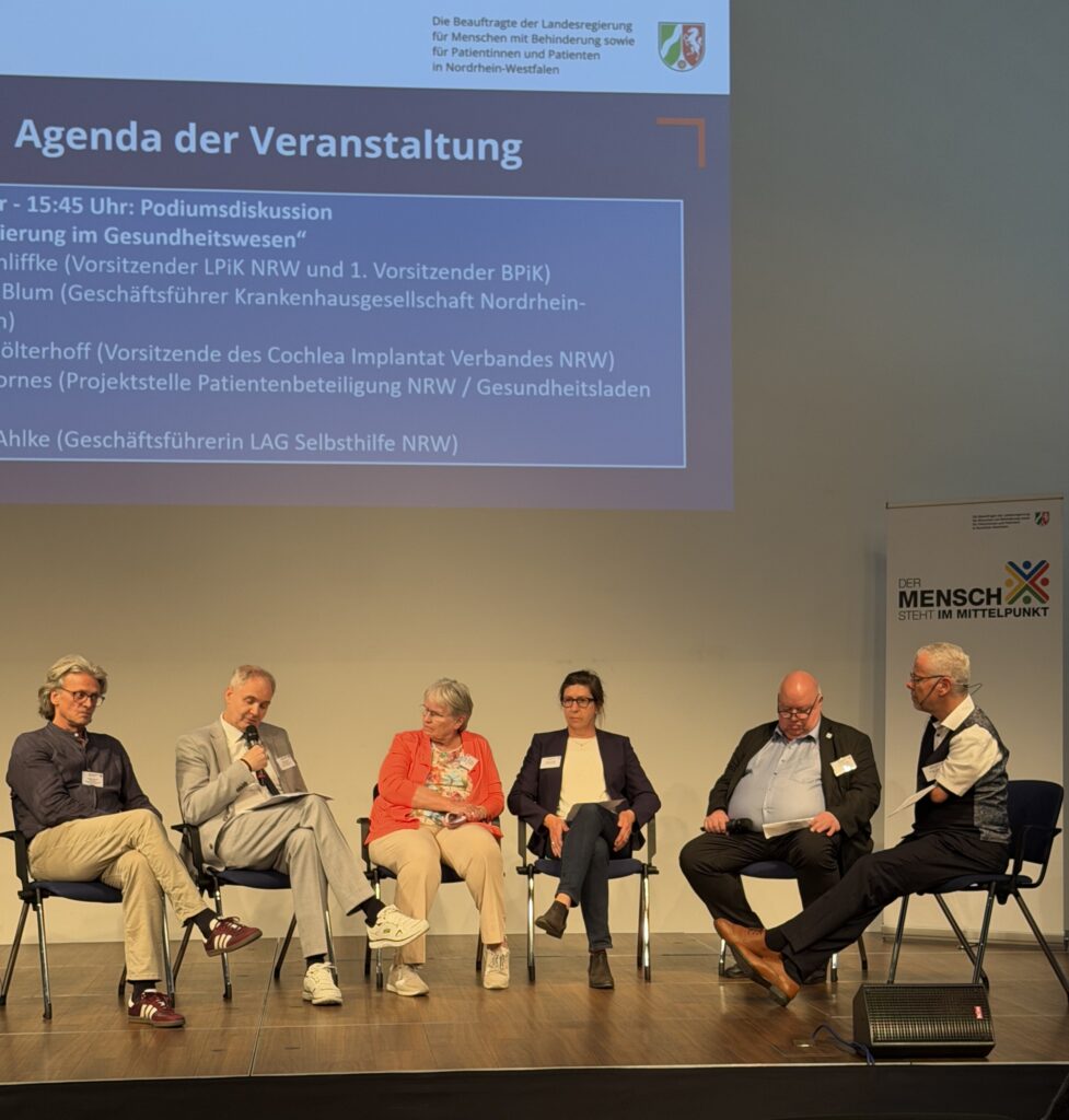 Auf einem Podium sitzen 6 Menschen auf Stühlen in einem Halbkreis - v. l. n. r.: Ein Mann mit mittellangen, grau gewellten Haaren und randloser Brille in dunkelblauem Hemd, beiger Hose und weinroten Sneakern. Ein älterer Herr mit kurzem Haar in grauem Anzug, der in das Mikro in seiner Hand spricht und dabei auf die Zettel auf seinem Knie schaut. Eine ältere Frau grauen, mittellangen Haaren in einer orangefarbenen Strickjacke über einem bunt gemustertem Shirt, die zum Sprecher neben ihr schaut. LAG-Geschäftsführerin Melanie Ahlke mit langen, dunklen, hochgesteckten Haaren und Hornbrille, in dunkler Hose und Blazer über einem hellem Shirt. Ein korpulenterer Mann mit Halbglatze, Brille und hellblauem Hemd unter einem dunklem Anzug. Und Moderator Rainer Schmidt im seitlichen Profil mit kurzen, grauen Haaren, Headset, Brille, dunkler Hose, Weste und weißem, kurzärmeligem Hemd, unter dem der linke Armstumpf hervorguckt. Im Hintergrundsind außerdem ein Roll-Up mit der Aufschrift „Der Mensch steht im Mittelpunkt“ sowie die Projektion einer Präsentation mit Ausschnitten der Agenda zur Podiumsdiskussion zu erahnen.