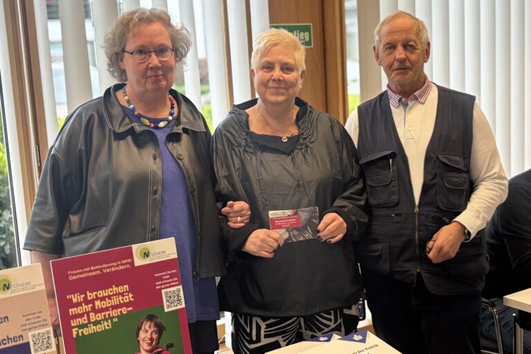 Monika Rosenbaum, steht hinter einem Tisch, auf dem sie verschiedene Broschüren und Poster des Netzwerk-Büros sowie des IZI-Projekts präsentiert – insbesondere „Wir brauchen mehr Mobilität und Barrierefreiheit“ und „Mitmachen bei der Politik an Ihrem Wohnort“. Sie hat helle helle, halblange, lockige Haare und trägt eine Brille, eine bunte Perlenkette sowie eine dunkle Jacke mit kurzen Ärmeln über einem blauen Shirt. Neben ihr lächeln eine ältere Dame und ein Herr im Rentenalter in die Kamera…
