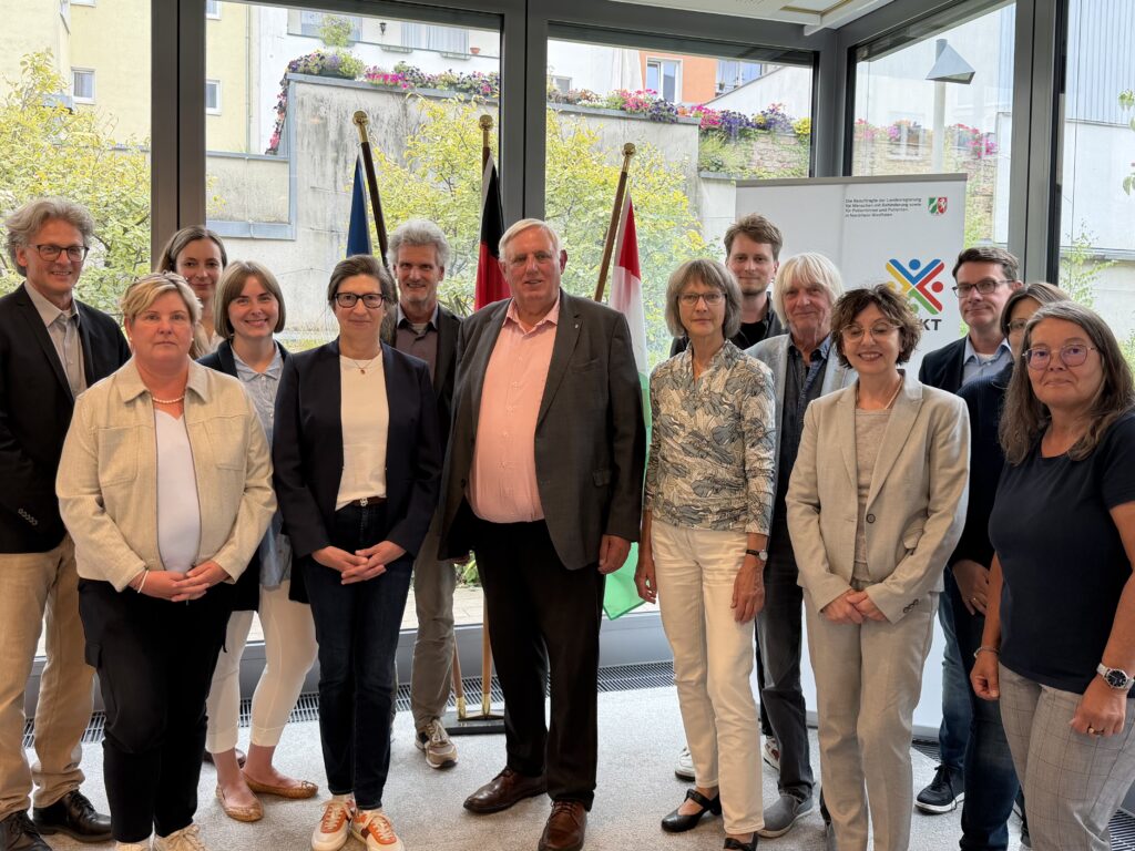14 Personen haben sich in einem verglasten Raum lose zum Gruppenbild vor drei Fahnen (Europa, Deutschland und NRW) sowie einem Roll-Up mit dem Logo der Beauftragten der Landesregierung für Menschen mit Behinderung sowie für Patientinnen und Patienten in Nordrhein-Westfalen aufgestellt. Claudia Middendorf (2. von links) hat kurze, blonde Haare, trägt eine hell Perlenkette zum biegen Blazer über weißem Shirt und hat die Hände gefaltet vor ihrer schwarzen Hose. LAG-Geschäftsführerin Melanie Ahlke (5. von links) lächelt mit langen, dunklen, zum Zopf nach hinten gekämmten Haaren und Hornbrille und trägt einen dunklen Blazer über weißem Shirt zu Jeans. NRW-Gesundheitsminister Karl-Josef Laumann (7. von links) guckt ebenfalls freundlich in die Kamera, hat graue, kurz gescheitelte Haare und trägt ein lachsfarbenes Hemd zu seinem grauen Anzug.