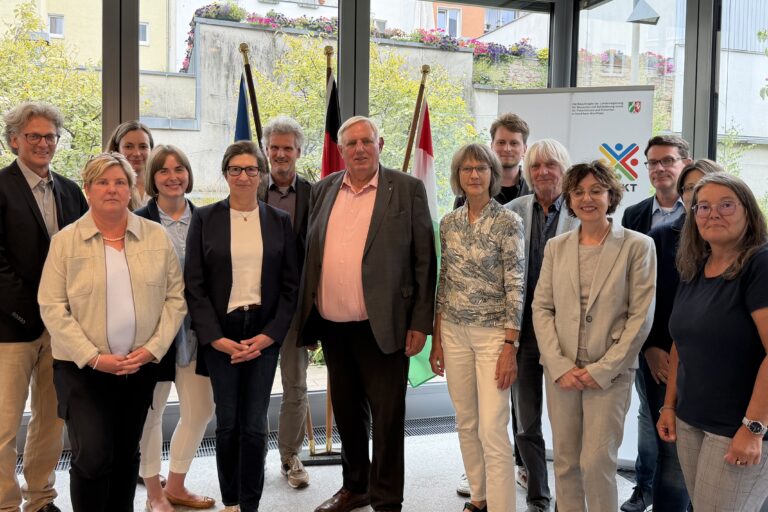 14 Personen haben sich in einem verglasten Raum lose zum Gruppenbild vor drei Fahnen (Europa, Deutschland und NRW) sowie einem Roll-Up mit dem Logo der Beauftragten der Landesregierung für Menschen mit Behinderung sowie für Patientinnen und Patienten in Nordrhein-Westfalen aufgestellt. Claudia Middendorf (2. von links) hat kurze, blonde Haare, trägt eine hell Perlenkette zum biegen Blazer über weißem Shirt und hat die Hände gefaltet vor ihrer schwarzen Hose. LAG-Geschäftsführerin Melanie Ahlke (5. von links) lächelt mit langen, dunklen, zum Zopf nach hinten gekämmten Haaren und Hornbrille und trägt einen dunklen Blazer über weißem Shirt zu Jeans. NRW-Gesundheitsminister Karl-Josef Laumann (7. von links) guckt ebenfalls freundlich in die Kamera, hat graue, kurz gescheitelte Haare und trägt ein lachsfarbenes Hemd zu seinem grauen Anzug.