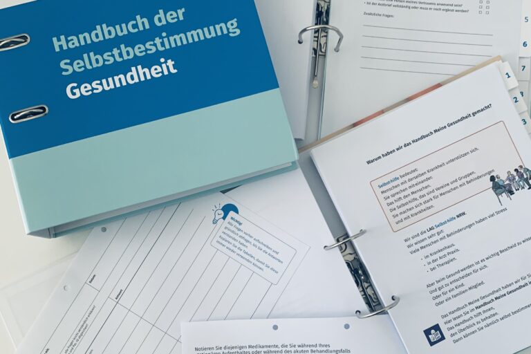 Neben dem Ringordner mit dem weiß-blau-mint gestreiften Cover des „Handbuch der Selbstbestimmung – Gesundheit“ der LAG Selbsthilfe NRW liegen zwei weitere aufgeschlagene Exemplare sowie zwei lose, ausgeheftete Blätter. Diese zeigen beispielsweise Checklisten für das Aufnahmegespräch, über eingenommene Medikamente während der Behandlung und Fragen zur Entlassung sowie eine Erklärung in Leichter Sprache, warum die Inhalte für Patient*innen wichtig sind.