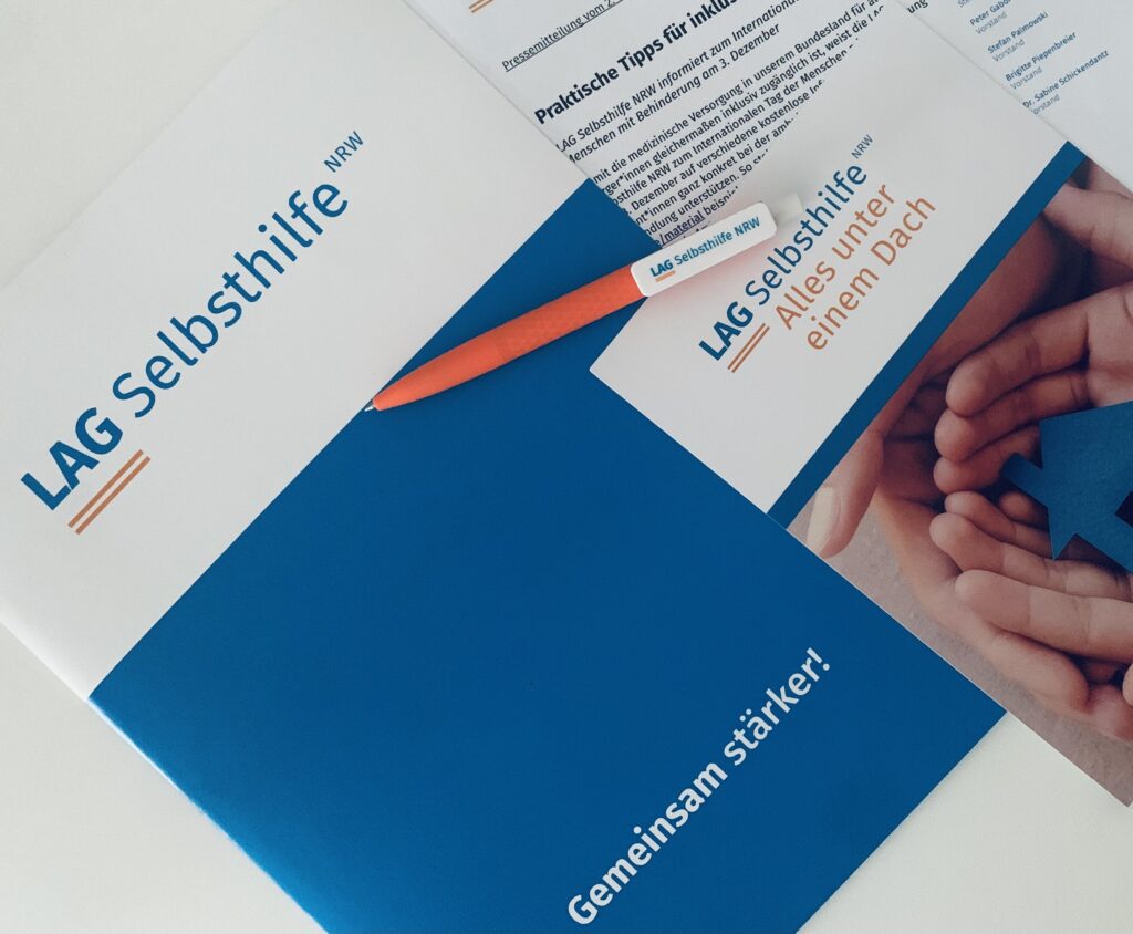 Das Symbolbild zeigt eine Dokumentenmappe im Corporate Design der LAG Selbsthilfe NRW mit dem Logo auf weißem Hintergrund und dem blau hinterlegten Claim „Gemeinsam stärker!“. Darauf liegen unser orangefarbener Kugelschreiber, der Imageflyer mit dem Bild eines gebastelten Hauses zwischen zwei Händen unter dem Schriftzug „Alle unter einem Dach“ sowie eine ausgedruckte Pressemitteilung.