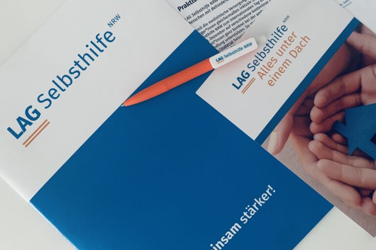 Das Symbolbild zeigt eine Dokumentenmappe im Corporate Design der LAG Selbsthilfe NRW mit dem Logo auf weißem Hintergrund und dem blau hinterlegten Claim „Gemeinsam stärker!“. Darauf liegen unser orangefarbener Kugelschreiber, der Imageflyer mit dem Bild eines gebastelten Hauses zwischen zwei Händen unter dem Schriftzug „Alle unter einem Dach“ sowie eine ausgedruckte Pressemitteilung.