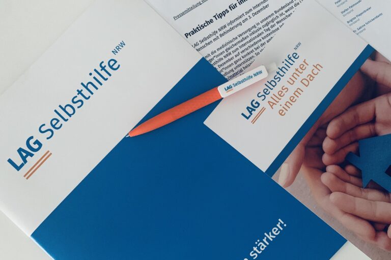 Das Symbolbild zeigt eine Dokumentenmappe im Corporate Design der LAG Selbsthilfe NRW mit dem Logo auf weißem Hintergrund und dem blau hinterlegten Claim „Gemeinsam stärker!“. Darauf liegen unser orangefarbener Kugelschreiber, der Imageflyer mit dem Bild eines gebastelten Hauses zwischen zwei Händen unter dem Schriftzug „Alle unter einem Dach“ sowie die ausgedruckte Pressemitteilung.