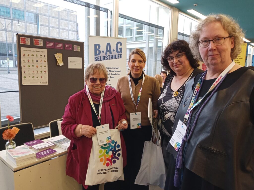 Vor einem Tisch mit Broschüren und Flyers, einer Stellwand mit angepinnten Postkarten sowie einem Roll-Up am Infostand der BAG Selbsthilfe stehen die vier Frauen und lächeln in die Kamera.