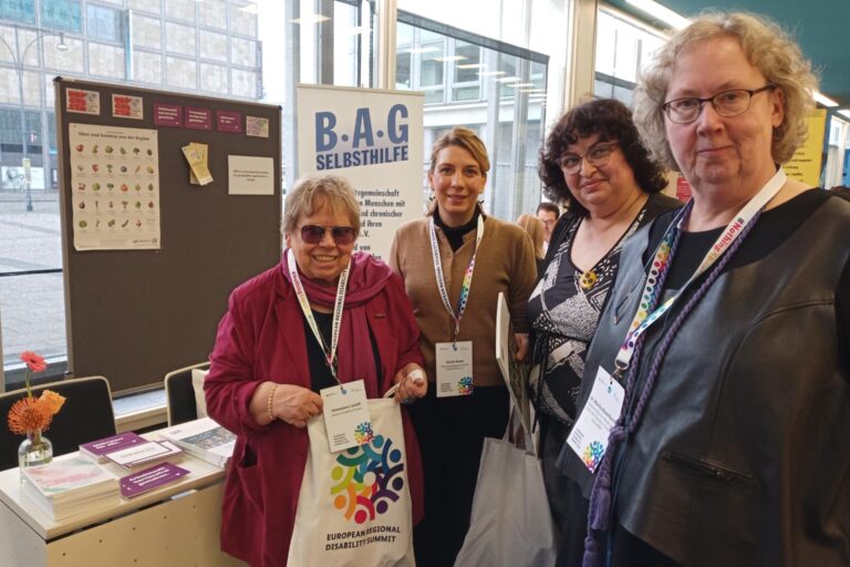Vor einem Tisch mit Broschüren und Flyers, einer Stellwand mit angepinnten Postkarten sowie einem Roll-Up am Infostand der BAG Selbsthilfe stehen die vier Frauen und lächeln in die Kamera.