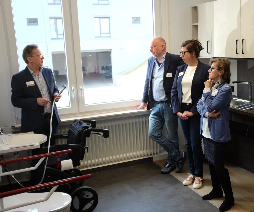 Vor einer anthrazitfarbenen Küchenzeile mit unterfahrbarer Spüle und hellen Hängeschränken stehen ein Besucher mittleren Alters mit leichtem Bart und Haarwuchs in dunkelblauem Jackett, hellblauem Hemd und Jeanshose, LAG-Geschäftsführerin Melanie Ahlke mit langen, brünetten, hochgesteckten Haaren, schwarz eingefasster Brille, in dunklem Blazer, hellem Shirt und Jeans sowie eine etwas kleinere Frau mit hellen, mittellangen Haaren, dunkler Brille und beige gemustertem Seidenschal in hellblauer Strickjacke, dunkel kariertem Rock und schwarzen Stiefeln. Sie schauen interessiert zu einem gegenüber stehenden Heern in dunkler Anzugsjacke und hell gemustertem Hemd, der einen abnehmbaren Wasserkopf neben Waschbecken und WC im Bad-Bereich des Ausstellungsraumes demonstriert.