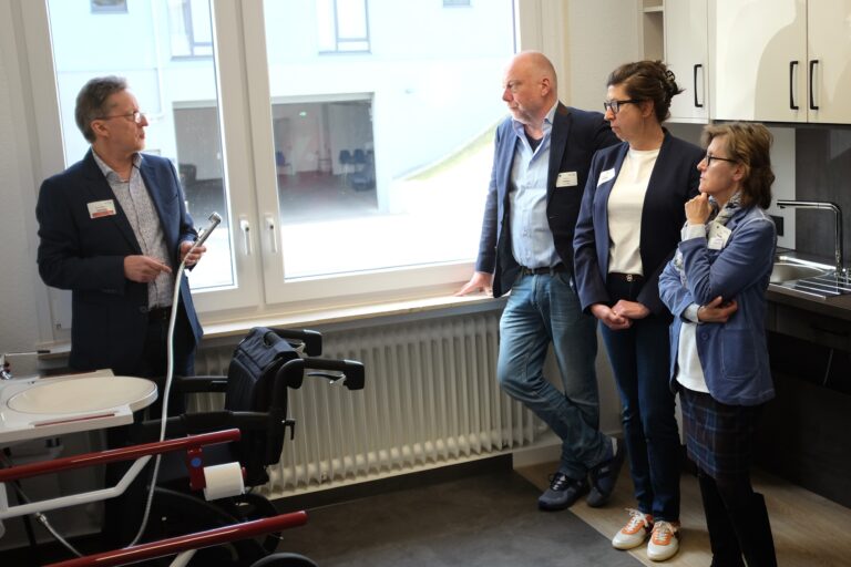 Vor einer anthrazitfarbenen Küchenzeile mit unterfahrbarer Spüle und hellen Hängeschränken stehen ein Besucher mittleren Alters mit leichtem Bart und Haarwuchs in dunkelblauem Jackett, hellblauem Hemd und Jeanshose, LAG-Geschäftsführerin Melanie Ahlke mit langen, brünetten, hochgesteckten Haaren, schwarz eingefasster Brille, in dunklem Blazer, hellem Shirt und Jeans sowie eine etwas kleinere Frau mit hellen, mittellangen Haaren, dunkler Brille und beige gemustertem Seidenschal in hellblauer Strickjacke, dunkel kariertem Rock und schwarzen Stiefeln. Sie schauen interessiert zu einem gegenüber stehenden Heern in dunkler Anzugsjacke und hell gemustertem Hemd, der einen abnehmbaren Wasserkopf neben Waschbecken und WC im Bad-Bereich des Ausstellungsraumes demonstriert.