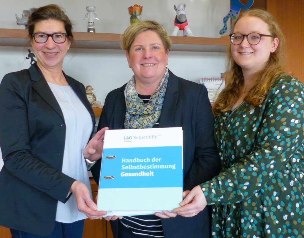 Melanie Ahlke, Lisa Handeck und Claudia Middendorf haben sich vor einem Büroregal zum Foto aufgestellt und halten lächelnd das „Handbuch der Selbstbestimmung – Gesundheit“ in die Kamera.