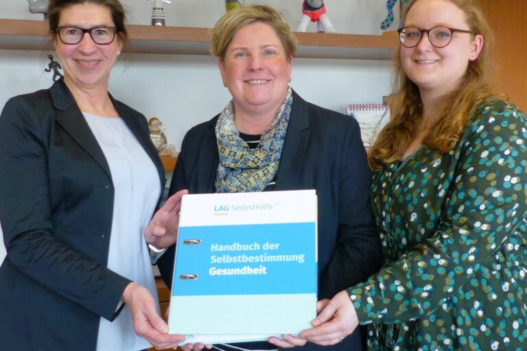 Melanie Ahlke, Lisa Handeck und Claudia Middendorf haben sich vor einem Büroregal zum Foto aufgestellt und halten lächelnd das „Handbuch der Selbstbestimmung – Gesundheit“ in die Kamera.