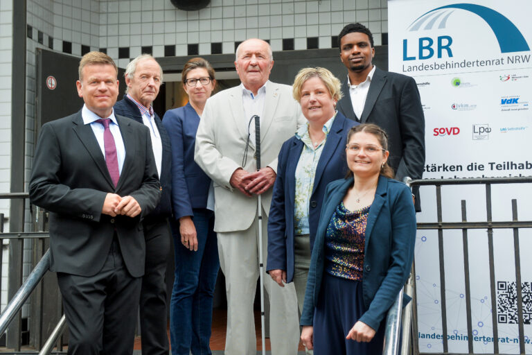 Neben einem Roll-Up, das die Logos das LBR NRW sowie seiner Mitgliederverbände (Lebenshilfe, Netzwerk Frauen und Mädchen mit Behinderung / chronischer Erkrankung NRW, LAG Werkstatträte NRW, Autismus Landesverband, Landesverband für Menschen mit Körper- und Mehrfachbehinderung NRW, VdK NRW, Interessenvertretung Selbstbestimmt Leben NRW, SoVD NRW, Landesverband Psychiatrie-Erfahrener NRW, LAG Selbsthilfe NRW) und einen QR-Code mit Link zur Internetseite (www.landesbehindertenrat-nrw.de) haben sich sieben Personen lächelnd zum Gruppenbild aufgestellt – von links nach rechts: Eine mittelalte Frau mit langen brünetten Haaren und randloser Brille in blauer Hose und Blazer mit glitzerndem Shirt darunter, ein junger Mann im Anzug mit dunklerer Haut, kurzen schwarzen Haaren und Kinnbart, eine Frau Ende 50 mit hellen halblangen Haaren in einem blauen Blazer mit hell gemusterter Bluse darunter, ein älterer Mann mit grauer Halbglatze in beigem Anzug und einem Langstock in den Händen, eine Frau mit nach hinten gekämmten langen dunklen Haaren und schwarzer Hornbrille in dunkelblauem Blazer und Jeans, ein Herr mit kurzen grauen Haaren in dunkler Jacke über einem blau-rot-weiß karierten Hemd und ein mittelalter Mann mit blonden kurzen Haaren in schwarzem Anzug, weißem Hemd und dunkelroter Krawatte.