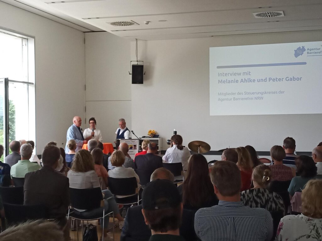 Der Blick geht von hinten über das Publikum in mehreren Stuhlreihen zur Bühne. Davor stehen Peter Gabor (mit weißen, kurzen Haaren, in hellblauer Anzugsjacke, den Blindenstock unter den Arm geklemmt) und Melanie Ahlke (mit Hornbrille und langen, dunklen, zu einem Dutt nach oben frisierten Haaren, in einem weißen Pullover, das Mikrofon in der Hand) von der LAG sowie Moderator Rainer Schmidt (mit graumelierter Kurzhaarfrisur, in einer blauen Weste über dem kurzen Hemd, aus dessen Ärmeln an beiden Seiten ein wenig die Stümpfe herausragen) an einem Stehtisch mit oranger Tischdecke links neben einem Rednerpult. Im Hintergrund ist die Präsentation an der Wand zu sehen, auf der unter dem Logo der Agentur Barrierefrei NRW steht: „Interview mit Melanie Ahlke und Peter Gabor – Mitglieder des Steuerungskreises der Agentur Barrierefrei NRW“