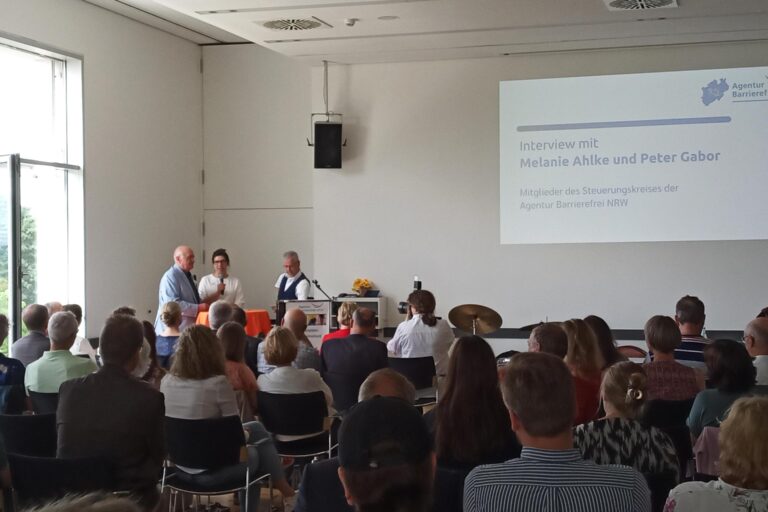 Der Blick geht von hinten über das Publikum in mehreren Stuhlreihen zur Bühne. Davor stehen Peter Gabor (mit weißen, kurzen Haaren, in hellblauer Anzugsjacke, den Blindenstock unter den Arm geklemmt) und Melanie Ahlke (mit Hornbrille und langen, dunklen, zu einem Dutt nach oben frisierten Haaren, in einem weißen Pullover, das Mikrofon in der Hand) von der LAG sowie Moderator Rainer Schmidt (mit graumelierter Kurzhaarfrisur, in einer blauen Weste über dem kurzen Hemd, aus dessen Ärmeln an beiden Seiten ein wenig die Stümpfe herausragen) an einem Stehtisch mit oranger Tischdecke links neben einem Rednerpult. Im Hintergrund ist die Präsentation an der Wand zu sehen, auf der unter dem Logo der Agentur Barrierefrei NRW steht: „Interview mit Melanie Ahlke und Peter Gabor – Mitglieder des Steuerungskreises der Agentur Barrierefrei NRW“