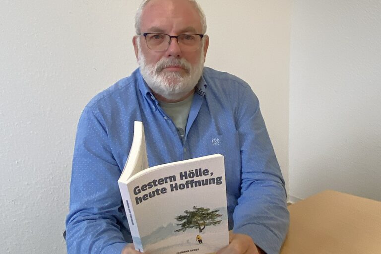 Ein älterer Herr mit grauem Vollbart und verbliebenem Haupthaar sowie dunkelrandiger Brille sitzt in einem hellblauen Hemd an einem Tisch. In seinen Hönden hält Clemens Sprey sein im Selbstverlag veröffentlichtes Buch „Gestern Hölle, heute Hoffnung“, das auf dem Cover einen grünen Baum vor der Kulisse einer grauen Berglandschaft zeigt.