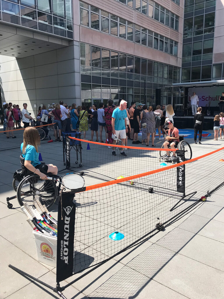 An einem hüfthoch aufgespannten Tennisnetz stehen sich zwei Kinder im Rollstuhl gegenüber. Der Junge im orangefarbenen T-Shirt und dunkler Cap legt sich gerade den Ball für den Aufschlag zurecht, den das Mädchen im blauen T-Shirt mit dem Schläger in der rechen Hand erwartet. Ein älterer Herr in blauem T-Shirt und kurzer Hose steht mit einem Schläger in der Hand daneben und gibt Tipps.