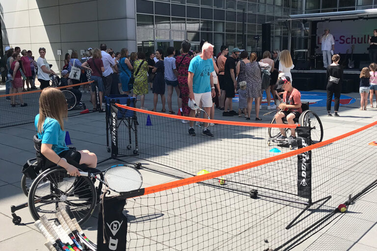 An einem hüfthoch aufgespannten Tennisnetz stehen sich zwei Kinder im Rollstuhl gegenüber. Der Junge im orangefarbenen T-Shirt und dunkler Cap legt sich gerade den Ball für den Aufschlag zurecht, den das Mädchen im blauen T-Shirt mit dem Schläger in der rechen Hand erwartet. Ein älterer Herr in blauem T-Shirt und kurzer Hose steht mit einem Schläger in der Hand daneben und gibt Tipps.