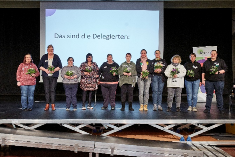 Die elf Delegierten haben sich auf einer Bühne nebeneinander zum Gruppenfoto aufgestellt, halten alle einen Blumenstrauß in den Händen und lächeln in die Kamera. Über ihren Köpfen ist der Schriftzug „Das sind die Delegierten:“ zu lesen, der auf eine Leinwand hinter ihnen projiziert wird.