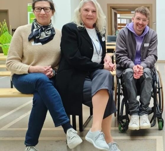 Dennis Sonne, ein etwa 40-jähriger Mann mit kurzen, lockigen Haaren, transparent eingefasster Brille und lila Kapuzen-Sweatshirt unter einer schicken, grauen Jacke, sitzt im Rollstuhl und lacht in die Kamera. Links neben ihm haben zwei mittelalte Frauen auf der Ecke einer Holzbank Platz genommen, die ebenfalls ein Lächeln auf den Lippen haben: Die Dame in der Mitte hat lange, helle Haare und ist mit einem schwarzen Blazer über einem weißen Shirt gekleidet, während LAG-Geschäftsführerin ihre dunklen Haare zu einem Dutt nach hinten gebunden hat und zum beigefarbenen Strickpullover ein grau-blau gemustertes Seidentuch sowie eine Hornbrille trägt.