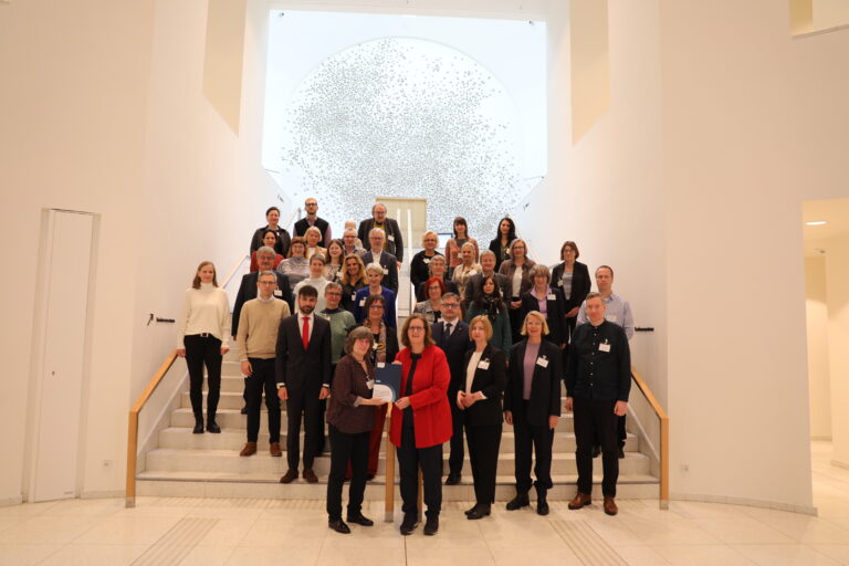 Das Foto zeigt rund 40 Mitglieder des Arbeitskreises Gewaltschutz für Menschen mit Behinderungen, die in mehreren Reihen auf einer Treppe stehen und in die Kamera schauen. Ganz vorne links übergab Frau Metternich (Starke.Frauen.Machen) den neu veröffentlichten Wegweiser am 14.11.2025 stellvertretend an die Parlamentarische Staatssekretärin Kerstin Griese (rechts).