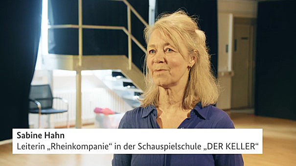 Der Screenshot aus einem Filmbeitrag zeigt eine mittelalte Frau mit langen blonden Haaren in dunkelblauer Bluse. Sie scheint in einer Interviewsituation vor einem Bühnenaufgang zu sprechen, der über eine etwa 8-stufige Treppe erreichbar ist und hinter einem dunklen Vorhang verborgen liegt, durch dessen Lücke ein helles Licht schimmert. Im unteren Bildbreich ist eine Bauchbinde mit folgenden Infos eingeblendet: Sabine Hahn - Leiterin „Rheinkompanie“ in der Schauspielschule „DER KELLER“