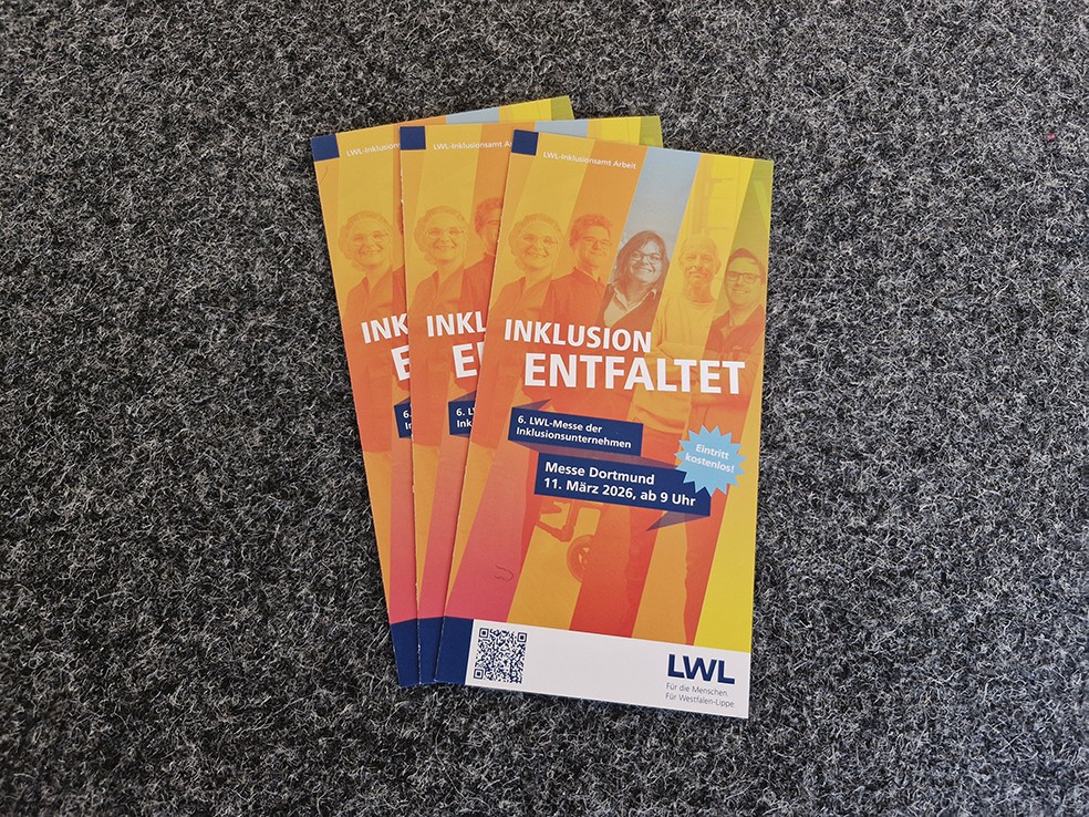 3 gleiche Flyer wurden leicht versetzt übereinander auf den einen grauen Teppichboden platziert. Sie zeigen die Fotos von 5 Mitarbeitenden unterschiedlichen Alters und Geschlechts mit verschiedenen Behinderungen, die jeweils in einem anderen Farbton hinterlegt sind. Über diesen „bunten Fächer“ aus Gelb, Orange, Blau, Gelb und Grün ist in Weiß der Titel „Inklusion entfaltet“ abgedruckt. Das Cover zeigt außerdem die weiteren Infos auf blauem Kontrast (6. LWL-Messe der Inklusionsunternehmen, Messe Dortmund, 11. März 2026, ab 9 Uhr, Eintritt kostenlos) sowie das Logo des LWL.