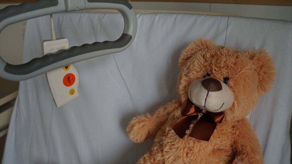 Symbolbild eines Krankenhausbettes, auf dessen Kopfkissen ein brauner Kuschelteddy mit roter Schleife um den Hals liegt.