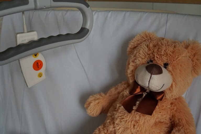 Symbolbild eines Krankenhausbettes, auf dessen Kopfkissen ein brauner Kuschelteddy mit roter Schleife um den Hals liegt.