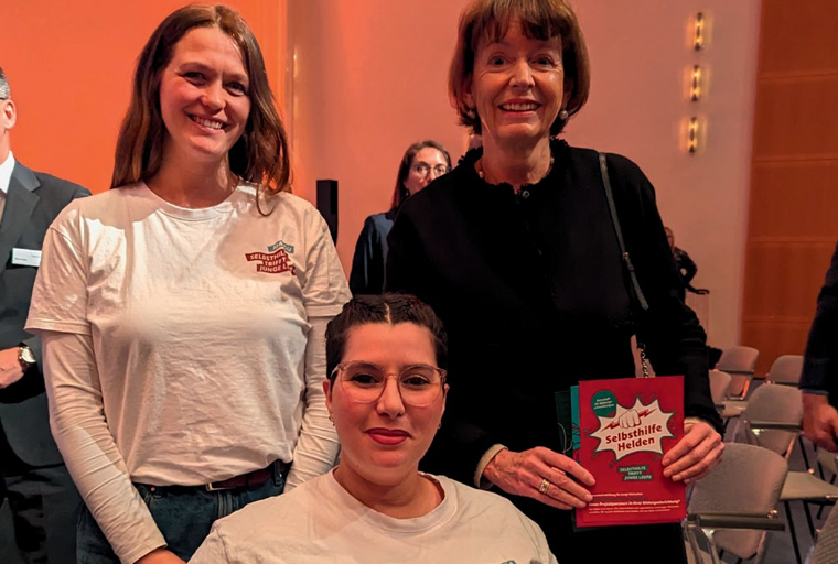 Malou Roy steht neben Frau Reker die einen Flyer der SelbsthilfeHelden in der Hand hält. Faiza Möwes sitzt in einem Rollstuhl vor den beiden, alle drei lächeln.