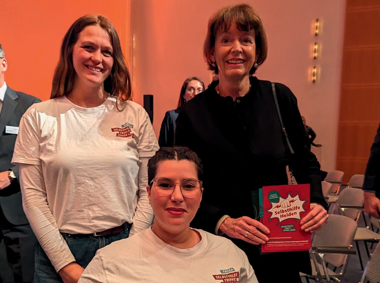Malou Roy steht neben Frau Reker die einen Flyer der SelbsthilfeHelden in der Hand hält. Faiza Möwes sitzt in einem Rollstuhl vor den beiden, alle drei lächeln.