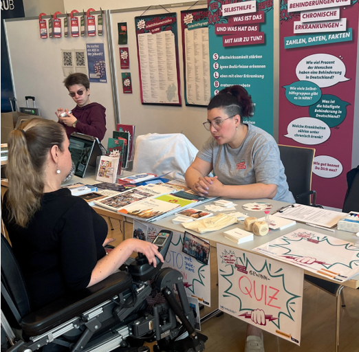 Faiza Möwes sitzt am Stand der SelbsthilfeHelden am Tisch und spricht mit Frau Kusal, der Beauftragten für die Belange der Studierenden mit Behinderungen und chronischen Erkrankungen. Im Hintergrund sind Roll-ups und weitere Materialien der SelbsthilfeHelden.