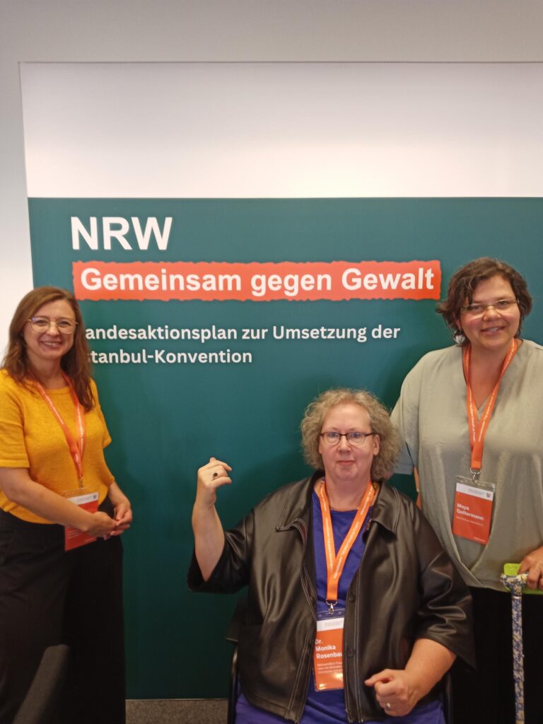 Monika Rosenbaum sitzt auf einem Stuhl mittig vor einem Aufsteller und zeigt mit dem Daumen der rechten Hand hinter sich auf die Aufschrift „NRW – Gemeinsam gegen Gewalt: Landesaktionsplan zur Umsetzung der Istanbul-Konvention“. Sie hat helle, lockige, halblange Haare, trägt eine fast randlose Brille und ein blaues Shirt unter einer schwarzen Lederjacke. An ihrem Hals hängt wie bei ihren Mitstreiterinnen ein oranges Namensschild mit gleichfarbigem Halteband. Leicht auf ihre Schulter gestützt steht rechts Maya Goltermann (mit Krückstock in der anderen linken Hand, Brille halblangen, dunklen Haaren, in einem hellgrünen Shirt und dunkler Hose) und links lächelt Julia Fischer-Suhr in die Kamera (mit langen brünetten Haaren und Brille, in gelbem T-Shirt und dunkler Hose).