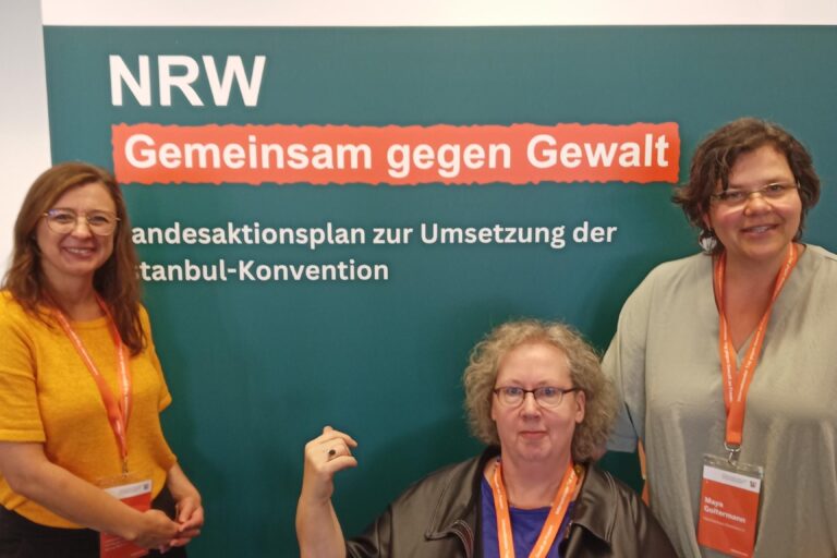 Monika Rosenbaum sitzt auf einem Stuhl mittig vor einem Aufsteller und zeigt mit dem Daumen der rechten Hand hinter sich auf die Aufschrift „NRW – Gemeinsam gegen Gewalt: Landesaktionsplan zur Umsetzung der Istanbul-Konvention“. Sie hat helle, lockige, halblange Haare, trägt eine fast randlose Brille und ein blaues Shirt unter einer schwarzen Lederjacke. An ihrem Hals hängt wie bei ihren Mitstreiterinnen ein oranges Namensschild mit gleichfarbigem Halteband. Leicht auf ihre Schulter gestützt steht rechts Maya Goltermann (mit Krückstock in der anderen linken Hand, Brille halblangen, dunklen Haaren, in einem hellgrünen Shirt und dunkler Hose) und links lächelt Julia Fischer-Suhr in die Kamera (mit langen brünetten Haaren und Brille, in gelbem T-Shirt und dunkler Hose).
