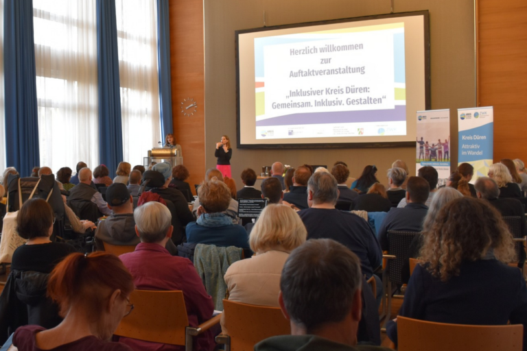 Blick auf die Bühne vom Ende des Veranstaltungsraums. Viele Teilnehmer*innen sitzen in Stuhlreihen vor der Bühne und schauen nach vorn.