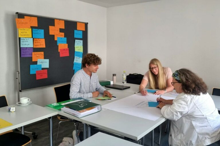 Die drei Inklusionsbeauftragten Niklas Beyer, Jennifer Schmandt und Emily Willkomm-Laufs sitzen gemeinsam an einem Tisch und blicken auf ein großes Blatt papier, das sie selbst beschrieben haben. Hinter ihnen steht eine Pinnwand mit bunten beschriebenen Zetteln.