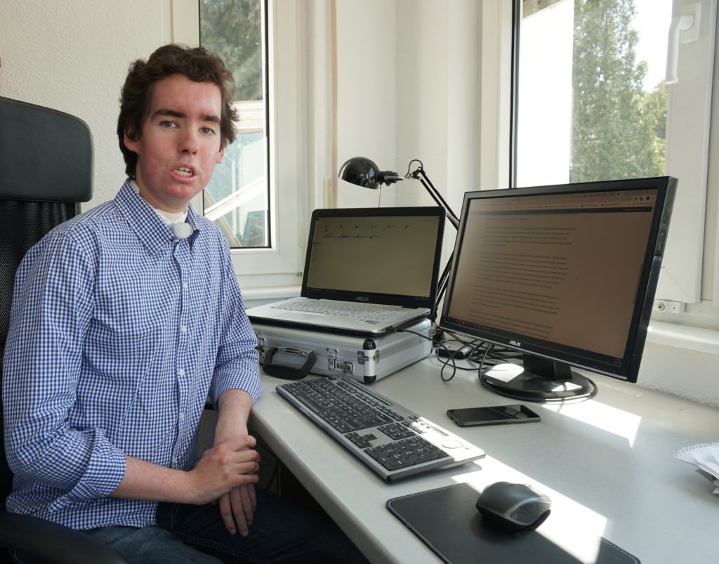 Hugo Schmidt sitzt aufrecht an einem Schreibtisch mit zwei Monitoren, einer Tastatur und einer Maus. Er Trägt ein blau-kariertes Hemd und blickt ernst in die Kamera.