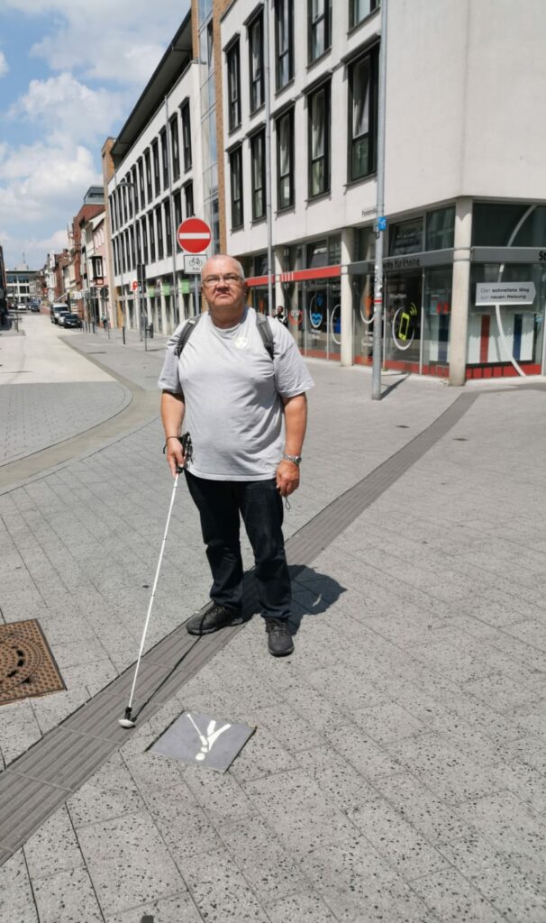 Caus Meier steht in der Innenstadt, er trägt ein hellgraues Tshirt, einen Rucksack und einen Langstock.