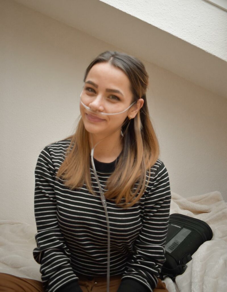 Sabrina blickt mit leicht angeschrägtem Kopf in die Kamera. Sie hat mittellange, braune Haare und trägt einen schwarz-weiß gestreiften Pullover. In der Nase befindet sich ein transparenter Schlauch, der zu einem Sauerstoffgerät führt.