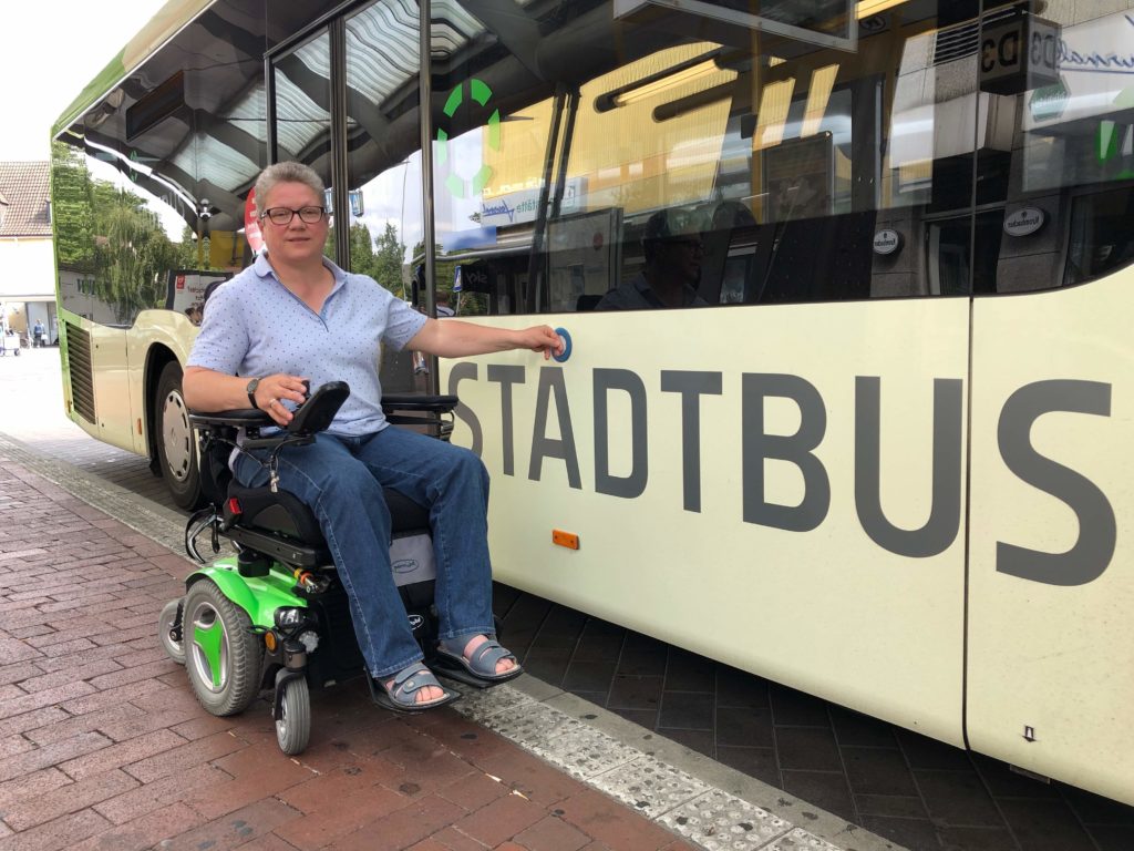 Frau Runte sitzt in ihrem Rollstuhl neben einem Stadtbus und drückt gerade auf den Knopf, der die Türen öffnet.