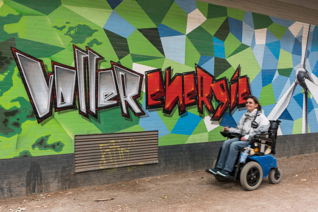 Silke Katzfuß fährt draußen in ihrem Rollstuhl durch. An der Wand befindet sich ein Graffitti. Es steht dort: Voller Energie.
