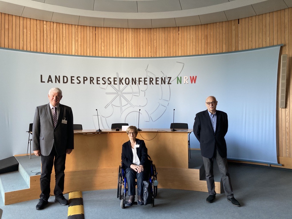 Franz Schrewe, Brigitte Piepenbreier und Horst Vöge blicken direkt in die Kamera. Hinter ihnen befindet sich ein großes Pult und eine helle Wand auf der steht: Landespressekonferenz NRW
