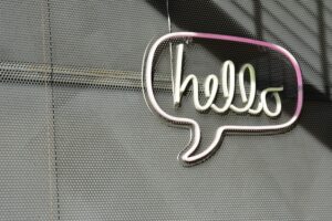 Eine graue Fassade an der ein Lichtbild in Form einer Sprechblase hängt. In ihr steht "Hello".