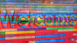 Eine Fassade aus bunten Holzleisten in blau-, grün-, rot- und lila-Tönen. In der Mitte ragt das Wort "Welcome" aus den Balken hervor. 